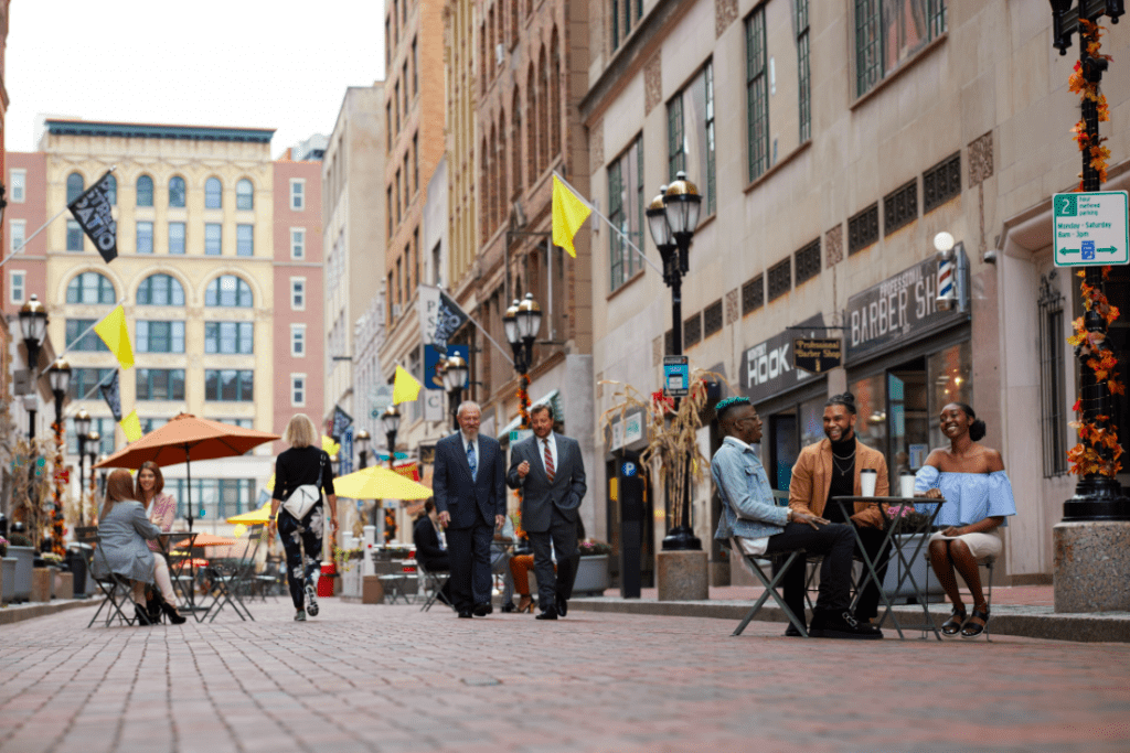 Millennial-aged people in groups of two and three are sitting around outdoor tables and two businessmen in suits are walking down the street.