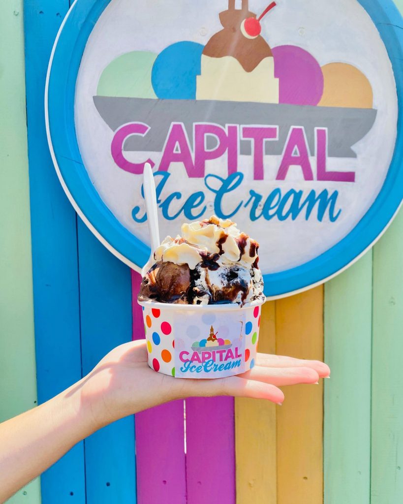 An ice cream sundae being held up by a single hand in front of a Capital Ice Cream sign