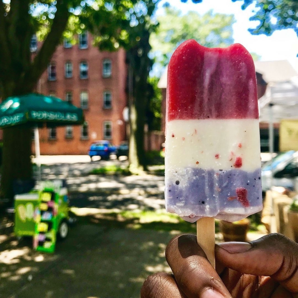 A hand holding up a red white and blue popsicle with trees and a building in the background