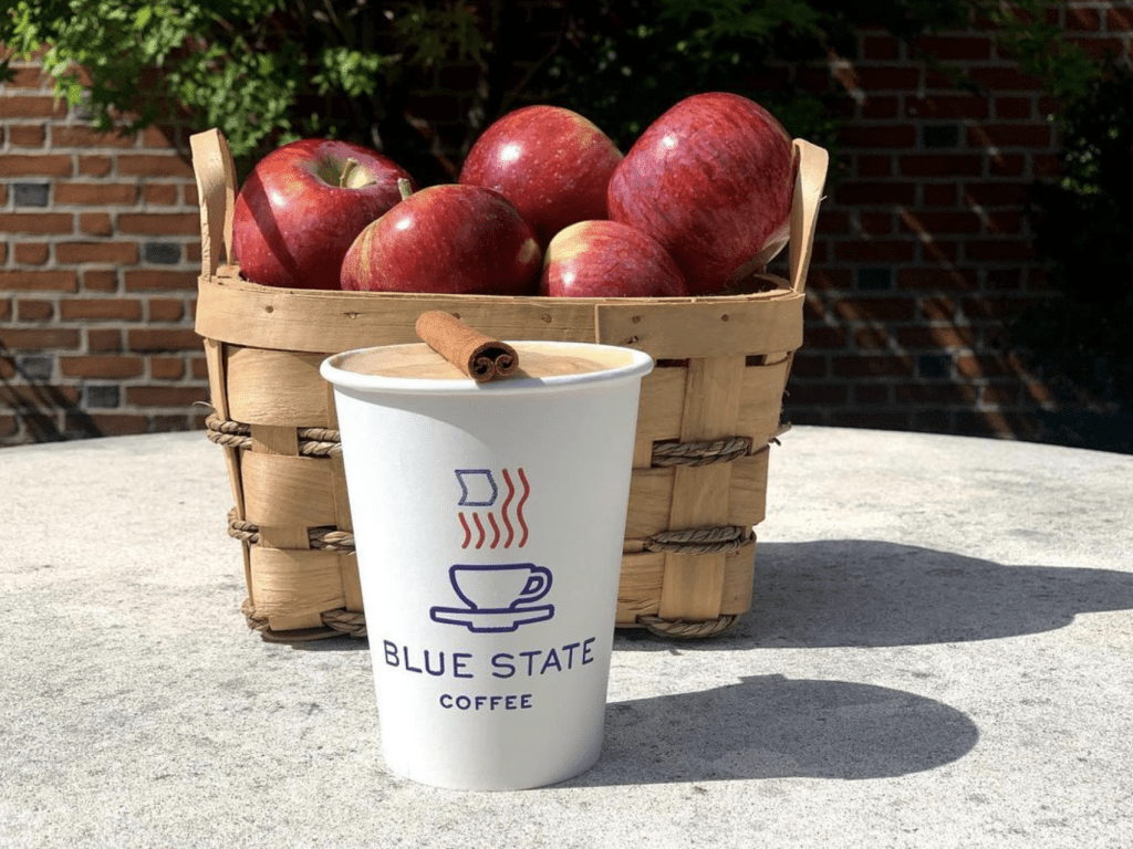 A chaider from Blue State Coffee in front of a bushel of apples.