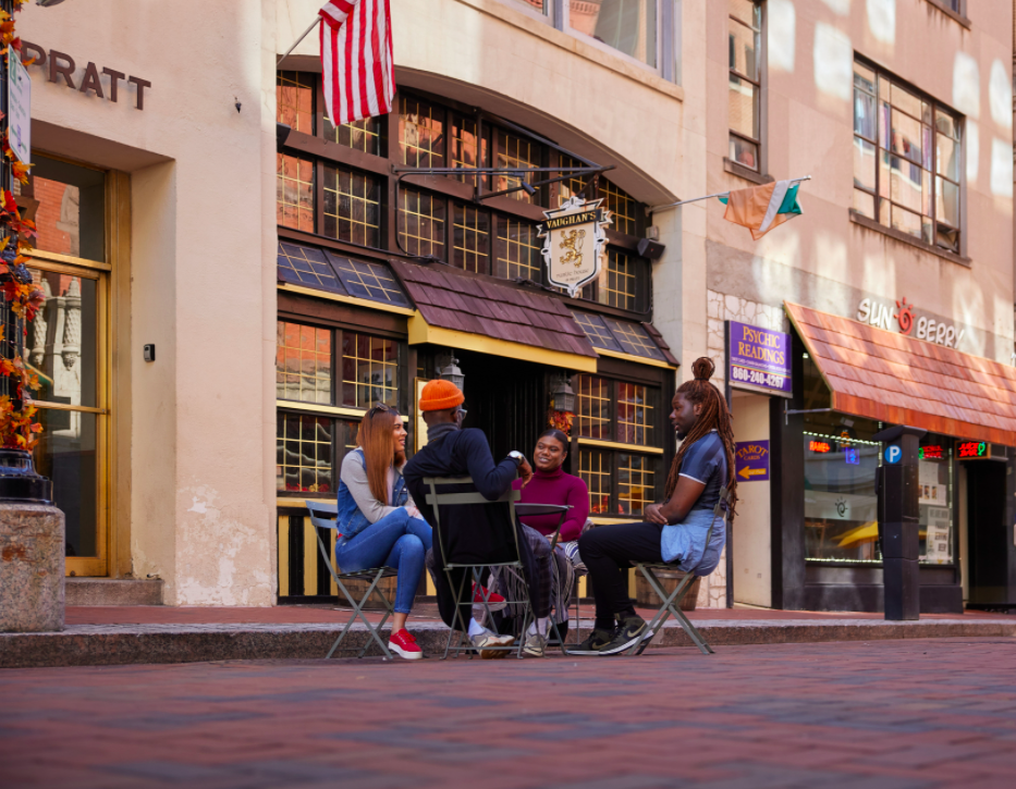 Four millennials sitting around an outdoor patio table on Pratt Street. The road is brick-lined and there are storefronts behind them.