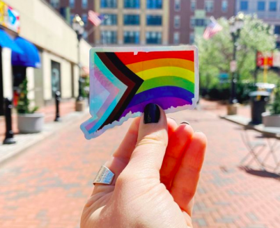 A Connecticut shaped sticker with the pride flag printed on it is held up on Pratt Street.