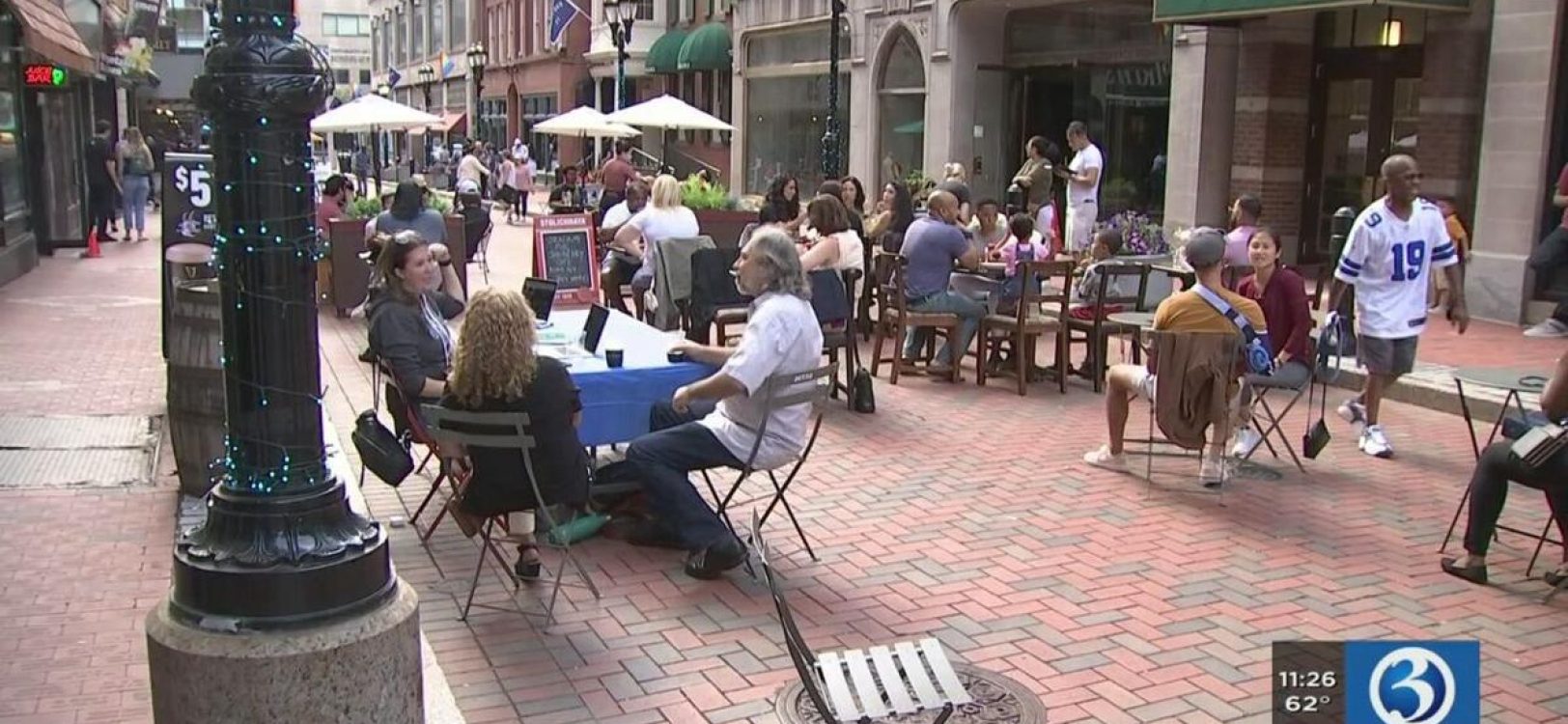 People sit at patio chairs around tables and stand and talk during the Pratt Street Summer Kickoff Party